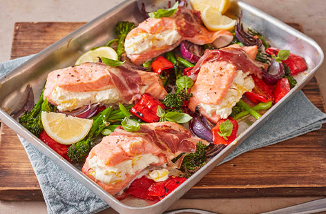 Cut pockets into salmon fillets and stuff with creamy soft cheese, wrap in Parma ham and roast with veggies for a super-easy traybake dinner.